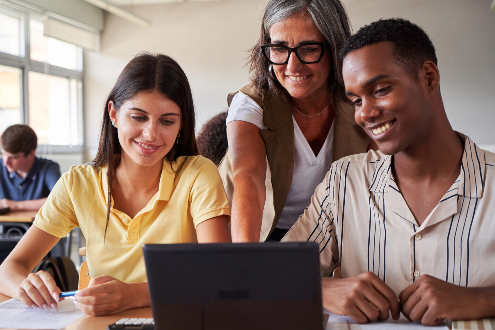 Smiling female teacher helping two multiracial college students using laptop to study in classroom. Image with copy space.