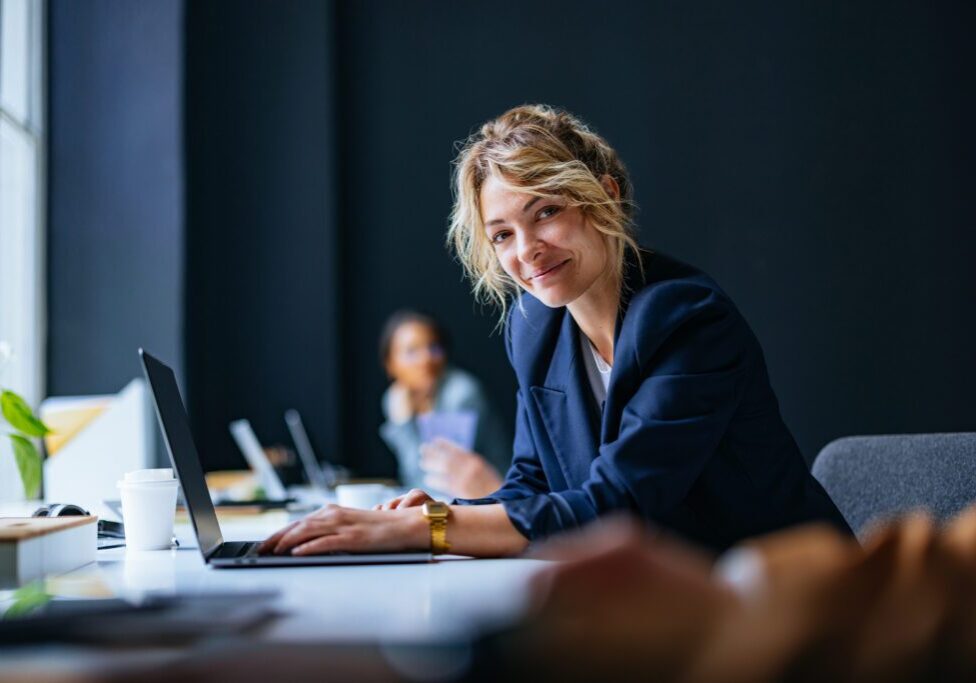Smiling businesswoman sitting at desk with laptop computer and looking at camera.