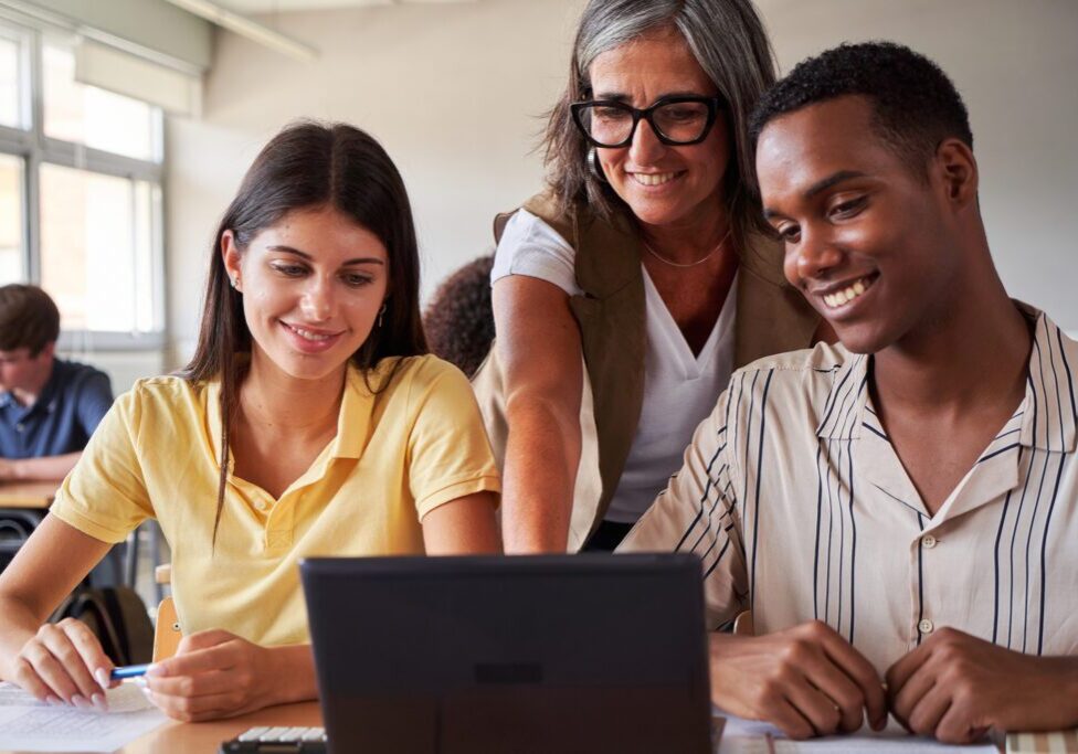 Smiling female teacher helping two multiracial college students using laptop to study in classroom. Image with copy space.